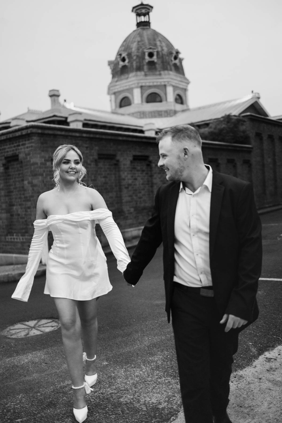A couple photographed by Meredith Photography in black and white. In the background Bathurst courthouse can be seen. The couple holding hands while looking at each other.